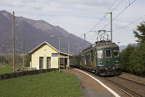 Einstöckiges Gebäude mit Satteldach neben dem Bahnsteig mit grünem Zug auf den Gleisen