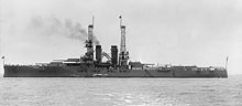 left side view of a large ship steaming through the water, with two boxy gun turrets in front and three in back, each with two guns.