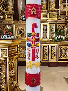 The Paschal candle with the cross and wax nails, the letters Alpha and Omega, and the year it was dedicated. As the cross reminds to a shepherd's crook, some sheep are depicted below to symbolise the flock. Ibaan Candela Paschalis closeup 2024-04-21.jpg