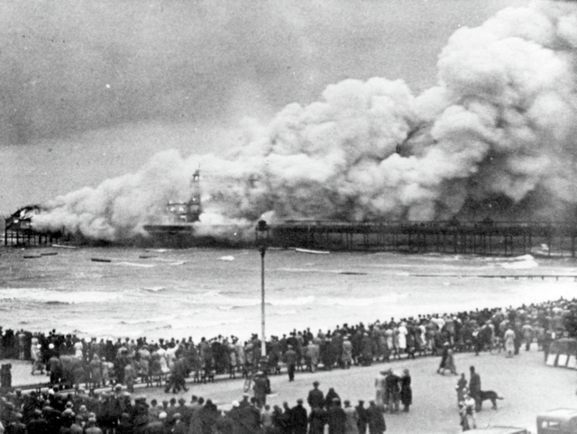 Black and white photograph of a pier almost entirely engulfed in smoke while a large crowd watches from the seafront