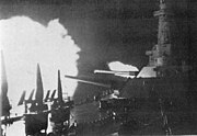 Two large gun turrets are trained to starboard, with the superstructure in the background. Flames are shooting out of one or more of the guns in both the nearer of the two turrets and an unseen gun astern.