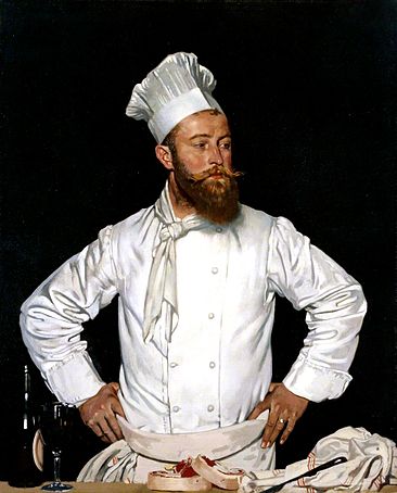 A chef with a bushy brown beard and waxed moustache wearing his white kitchen attire and a tall chef