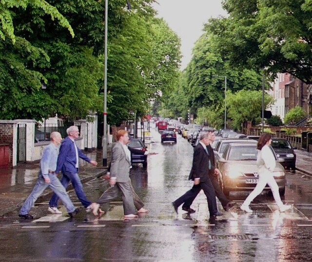 Imitating the famous cover of the Beatles album Abbey Road (1969), on which the band members cross the road in front of the Abbey Road Studios in a ro