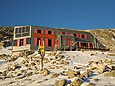 Eine moderne, zweigeschossige Berghütte mit Solarpanelen an einem Felshang, im Vordergrund Steine, Schnee und ein Wegweiser