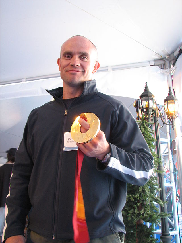 A hair-shaved man smiles at the camera while holding in his left hand a golden medal. He wears a dark sports jacket with a white stripe along the arm length, and is inside a large reception tent with a small street lamp and a pine tree behind.