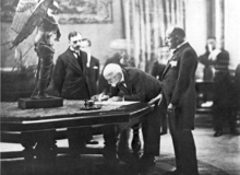 Un homme chauve penché sur une table de marbre signe un document tandis que deux hommes l'observent.