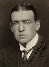 Man with hair centre-parted, wearing high white collar with tie, and a dark jacket. His facial expression is serious