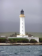 A tall white lighthouse with a brown stripe around the parapet and dark coloured lantern sit on a rocky shore. A white wall obscures the lower floor of grey stone buildings gathered around its base.