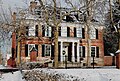 Red brick building with snow on the dark roof. In front of the building, there are trees and snow on the ground.