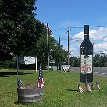 A grassy field along a road decorated with a black cardboard wine bottle and a handful of small flags, with a white wooden sign and flagpole in the background.