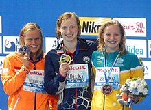 Die drei Schwimmerinnen posieren nach der Siegerehrung mit ihren Medaillen, die an einem blauen Band hängen. Die Sportlerinnen tragen ihre Trainingsjacke, an dem ihr Name auf Papier befestigt ist. Ashwood hält in ihrer linken Hand einen Blumenstrauß und das Maskottchen der WM.
