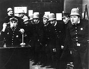 Left:Ford Sterling as Keystone Cops Police chief [seated}; 4th from right AL St John; 3th from right: Hank Mann;  in In the Clutches of the Gang (1914)