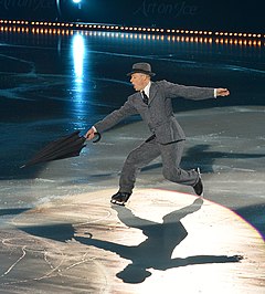 A man wearing a gray suit and fedora and holding an umbrella skates on ice