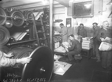 Photographie en noir et blanc d'hommes moustachus tenus des journaux papiers dans leurs mains en bout de chaîne de leur fabrication.