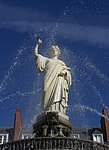 Statue blanche, sur fond de ciel bleu, encadrée par des jets d'eau