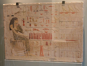 A flat limestone block with a painted, carved raised-relief of woman in spotted linen cloth, seated near table with food items. Painted hieroglyphs decorate the rest of the surface.