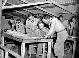 Circa otto uomini in uniforme militare che si sporgono sui fogli sulle scrivanie