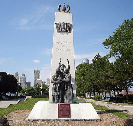 An Underground Railroad to Freedom monument, facing north-northwest from up Windsor's Civic Esplanade with the Detroit skyline distant across the river, from Pitt Street East, between Goyeau Street and McDougall Street, by the casino at a corner of The Colosseum.