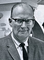 Photographie en noir et blanc d'un homme chauve portant un costume-cravate et des lunettes.