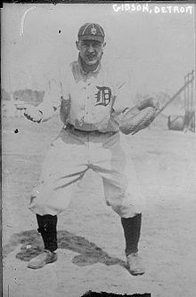 A man in a white baseball uniform with a dark "D" on the front, a dark cap with a white "D", and high socks stands with his arms apart awaiting a throw–his catcher's mitt.