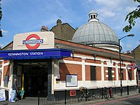 A red-bricked building with a rectangular, dark blue sign reading "KENNINGTON STATION" in white letters all under a light blue sky