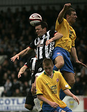 A player wearing a striped shirt, with his head close to the ball. Two players in yellow shirts are in very close proximity; one is jumping and making physical contact with him.