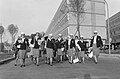 Un groupe de femmes vêtues de robes à mi-mollet et de vestes marche dans la rue. En fond, une grande barre d'immeubles et quelques arbres. La photo est en noir et blanc.