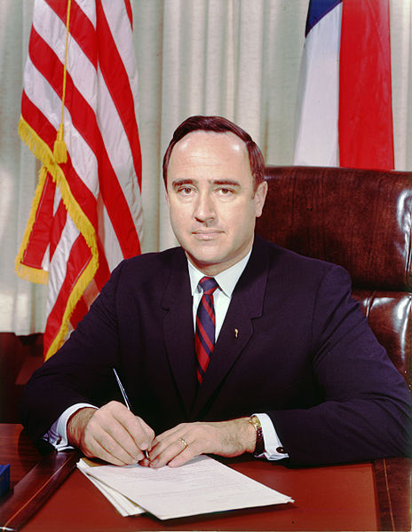 Governor Robert Walter Scott seated in his office. Two flags are behind him, and he is facing the camera with a pen in hand.