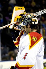 Un gardien de but de hockey sur glace enlevant son masque.  Il porte un maillot blanc et orange avec le numéro "1" sur son coude.