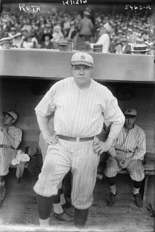 A man poses with his arms on his hips. He is wearing a striped jersey and pants, along with a baseball cap that has a logo with an interlocking "N" and "Y". Behind him, men wearing similar jerseys are sitting on a bench.