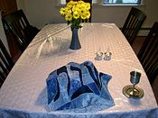The Shabbat table is set: two covered challot, a kiddush cup, two candles, and flowers. Shabbat table setting.jpg