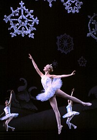 Three ballerinas dressed in white, dancing in a stage setting featuring giant snowflakes.