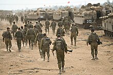 Men in desert-brown fatigues walk towards rows of tanks. All of them have backpacks, and some have firearms.