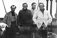 Four menwearing dishevelled clothing and worn expressions face the camera. The head of a dog is just visible on the left.