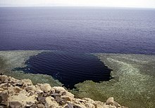 View of the coastal waters from the top of a hill, showing an approximately circular hole in the shallow coastal reef tangent to the deeper water offshore.