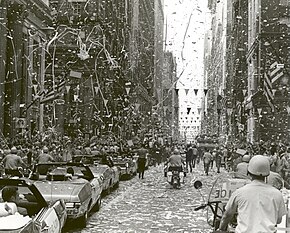 A city street among tall buildings with a procession of cars between bystanders, showered by shredded paper
