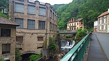 Foto reciente de la antigua fábrica Creux de l'Enfer tomada desde la orilla opuesta.  A la derecha la avenida, en el centro la Durolle con su cascada bajo un puente moderno, a la izquierda la fachada de la fábrica en 3 niveles.  La fábrica está construida en parte sobre bóvedas sobre el agua, el revestimiento de las paredes se está desmoronando y revela las piedras de pizarra en algunos lugares, todavía distinguimos un poco el nombre pintado en rojo.