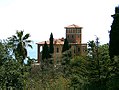 Vista del palacio de Hanbury, con su torre, en medio de un parque botánico lujoso. Cuatro cipréses y en el fondo, el mar azul. Una palmera.