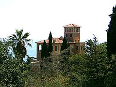 Vue du palais de Hanbury avec sa tour, au milieu d'un parc botanique luxuriant. Quatre cyprès et dans le fond, la mer bleue. Un palmier.