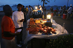 Caterers display food items for sale in the park. The Indian Ocean can be seen in the background. Forodhani park food stand.jpg