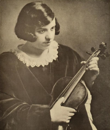 A teenaged girl with fair skin and dark hair, cut in a bob, wearing a dark dress with a lace collar detail; seated, holding a violin
