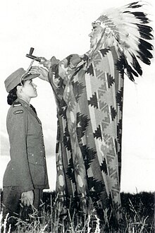 Photo de Mary Greyeyes agenouillée sur le sol tandis qu'un homme en costume traditionnel du chef cri des plaines agite une main sur son front