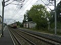 A train approaches the station. The former platform is on the right. The new platform is on the other side of the level crossing.