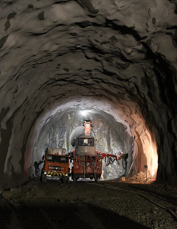 A tunnel under construction by conventional excavation methods with a pilot tunnel through tunnel face and a drill jumbo positioned near the face. Roc