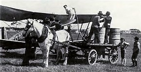 Photographie en noir et blanc représentant un avion de chasse de la Seconde Guerre mondiale au sol, ravitaillé avec des barils de carburants traînés par un cheval. Le pilote effectue l'opération, aidé par deux hommes.