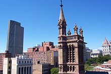 Eine Nahaufnahme des oberen Teils des Kirchturms mit einem Stadtbild dahinter. Links befindet sich ein hohes modernistisches Gebäude. Auf der rechten Seite befindet sich ein Turm mit einem roten Spitzdach