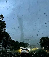 Cono centrale di tornado circondato da detriti volanti all'orizzonte.  Sotto l'orizzonte c'è una strada con veicoli fermi.  Sta piovendo.