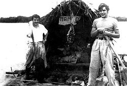 black and white photograph of two men on a raft, fitted with a large hut. The far bank of the river is visible in the far distance