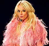 A blond woman in a red jacket with black fur on her shoulder. She is singing into a microphone.