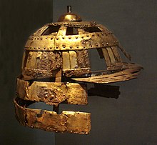 Foto colorida de um capacete de soldado feito de lâminas de ferro revestidas de cobre, sobre um fundo escuro.